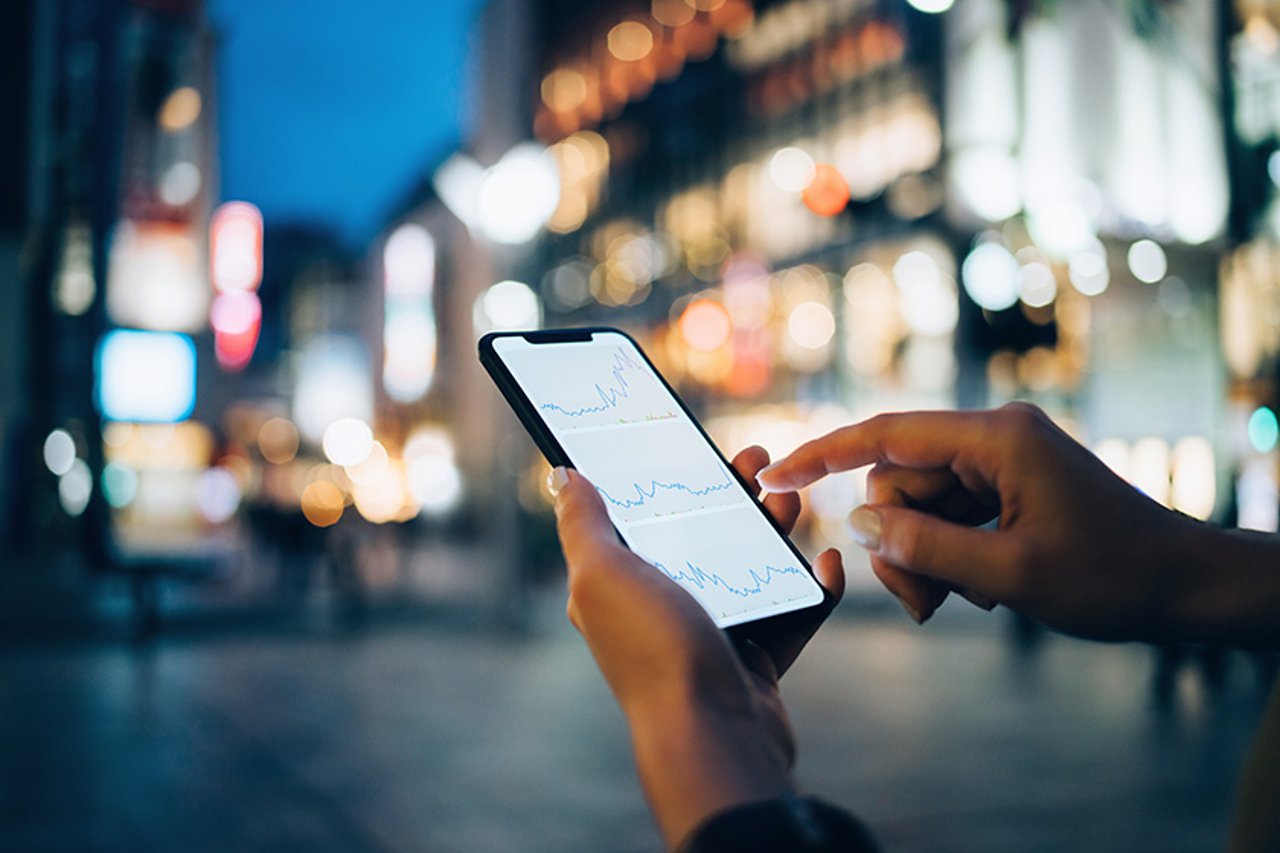 Les sociétés de services financiers novatrices utilisent des API de communication pour transformer l'expérience client, séduisant des clients parfois hésitants grâce à l'authentification en temps réel ainsi qu'aux échanges vidéo, vocaux et par message. Businesswoman reading financial trading data on smartphone in downtown city street against illuminated urban skyscrapers